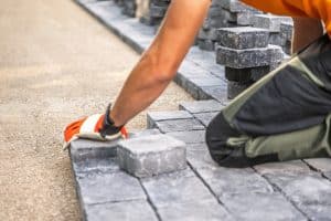 A worker wearing gloves installs gray interlocking pavers for a hardscaping project.