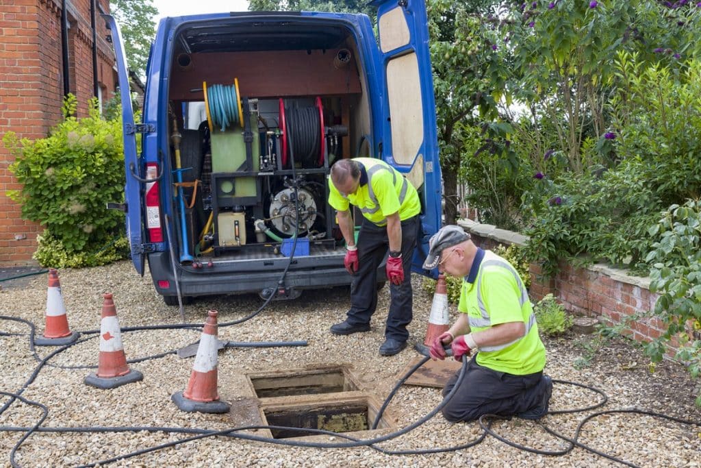 Drain cleaning workers using specialized equipment to clear a blocked underground drain outside a residential property.