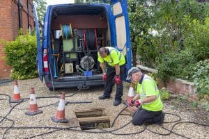 Drain cleaning workers using specialized equipment to clear a blocked underground drain outside a residential property.
